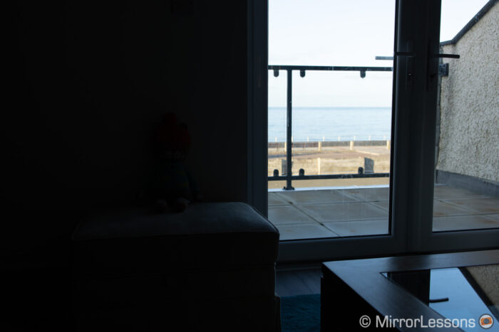 living room with window on the right showing outdoor balcony and sea in the background. The image is exposed for the outside (the sea) so the interior is very dark.