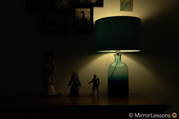 one japanese doll and two final fantasy action figures next to a lamp, on a chest of drawer. The image is visibly underexposed.